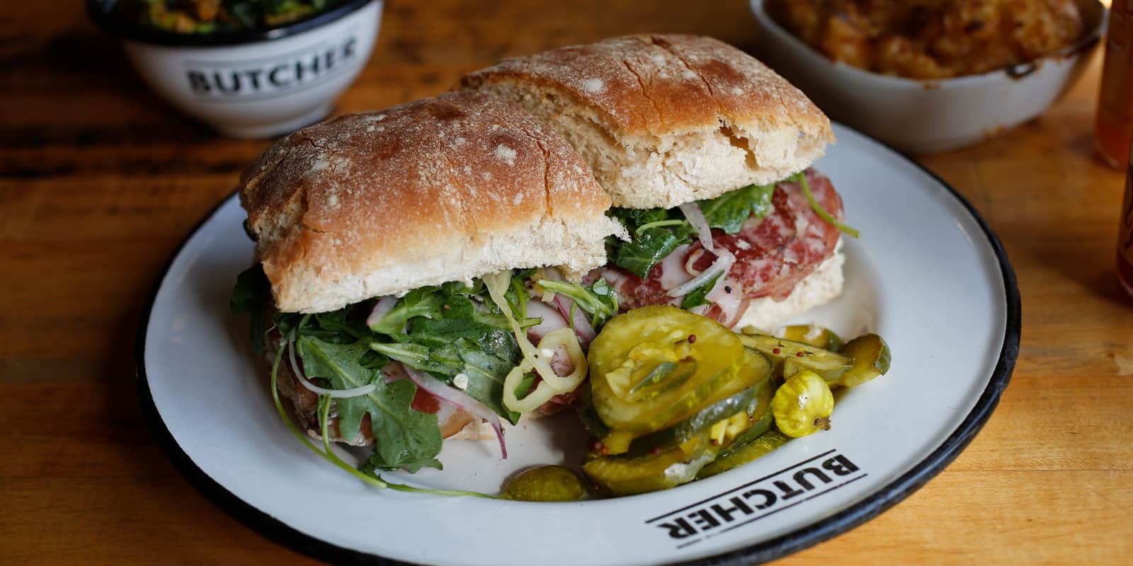 A dressed meat sandwich served on a plate next to sliced pickles.